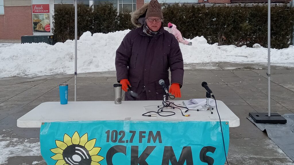 A man wearing a thick winter coat and hat, and bright orange gloves at table holding two microphones, and a banner that reads "102.7 FM CKMS"