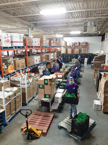 A picture of the food bank warehouse. Boxes and shelving and skids, and hand-carts, fluorescent lights, and even a few volunteers are in this busy colourful picture.