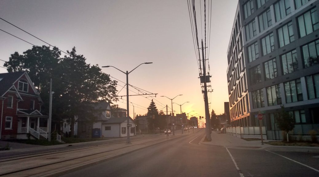 The sun sets over a 5-storey residential building on the right of the image. Cars in the distance drive east beside the LRT line.