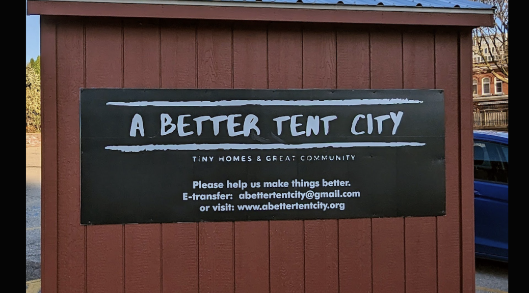 The side of a brown clapboard tiny home, measuring approximately 10 by 10. On the brown windowless side is a black banner that reads 'A Better Tent City'. These words are bordered top and bottom by white lines that are designed to look worn. Underneath in a smaller font are the words "Tiny Homes and Great Community. Please help us make things better.