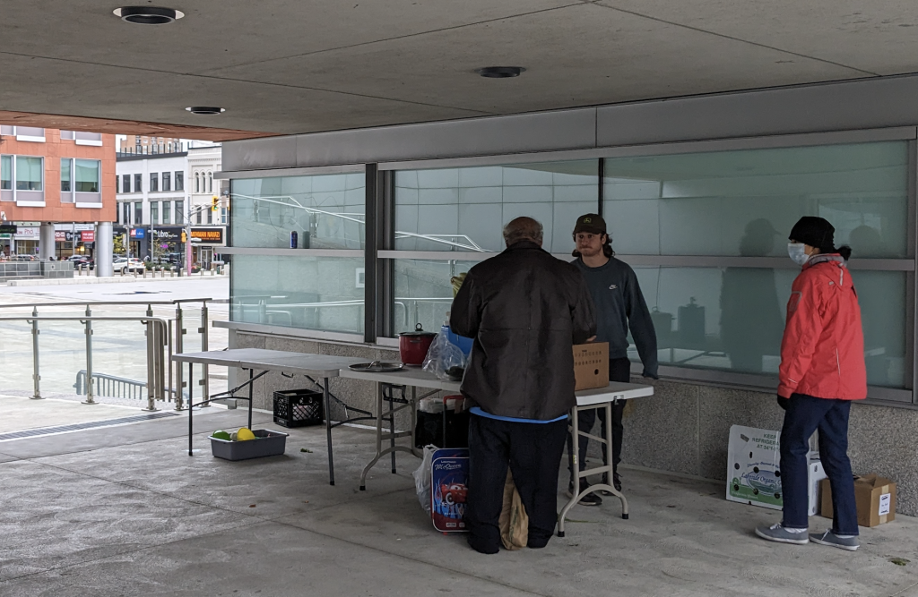 A person receives food from the Food Not Bombs table set up outside Kitchener City Hall. One person serves the food and another helps with the distribution.