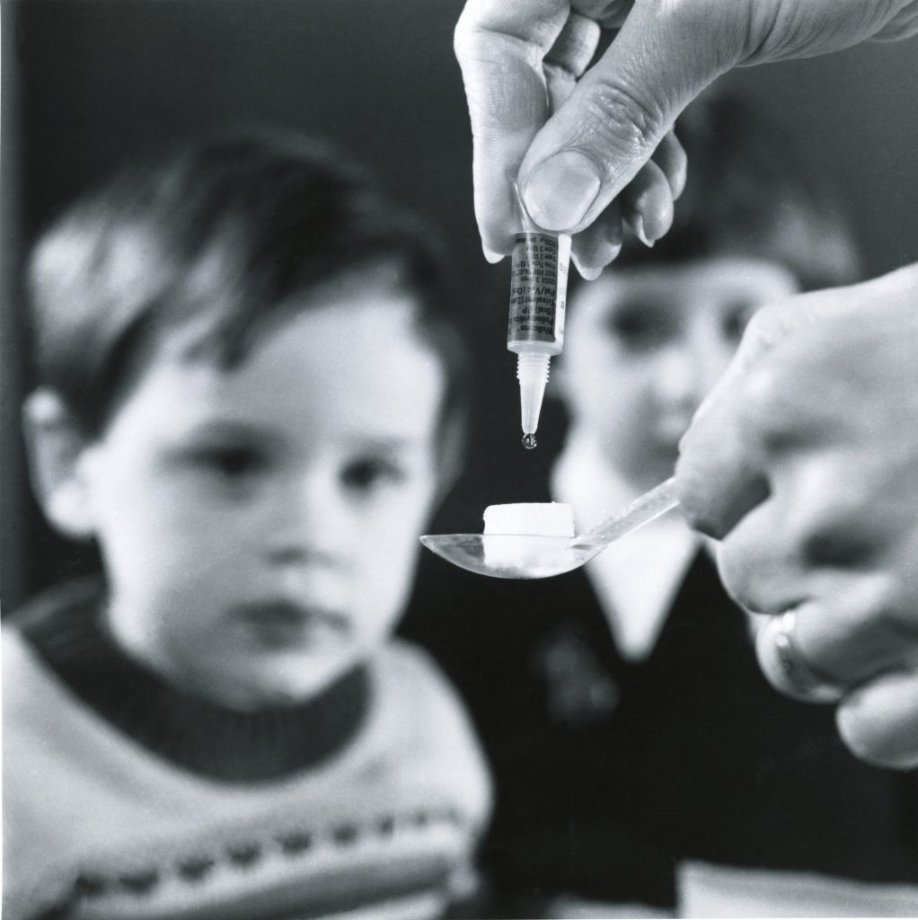 A black and white photo of two children sit unfocused in the background, staring at some polio vaccine being dripped from a dropper on a sugar lump which is being held on a spoon which is being held by a hand.