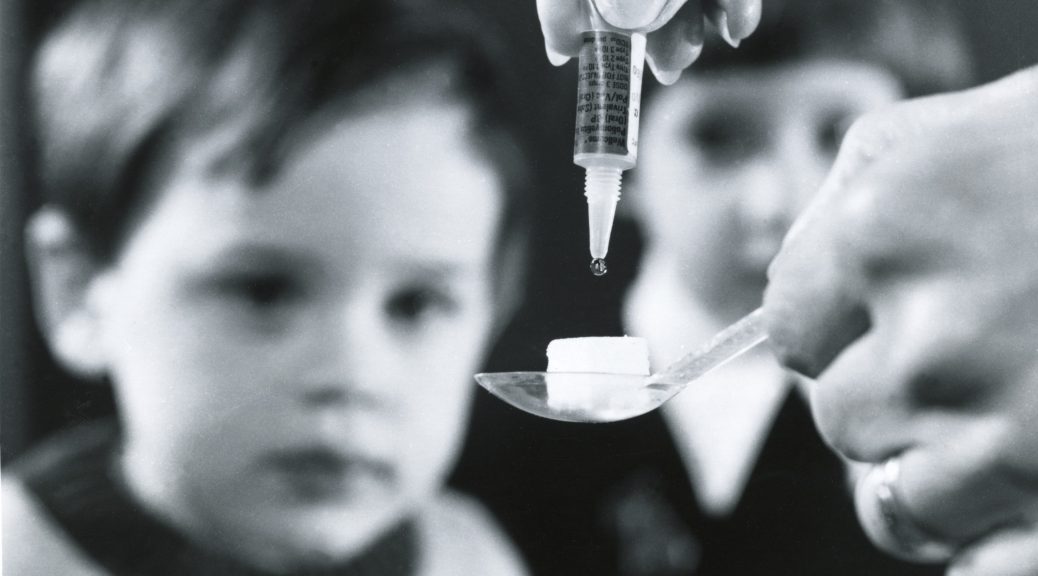 A black and white photo of two children sit unfocused in the background, staring at some polio vaccine being dripped from a dropper on a sugar lump which is being held on a spoon which is being held by a hand.