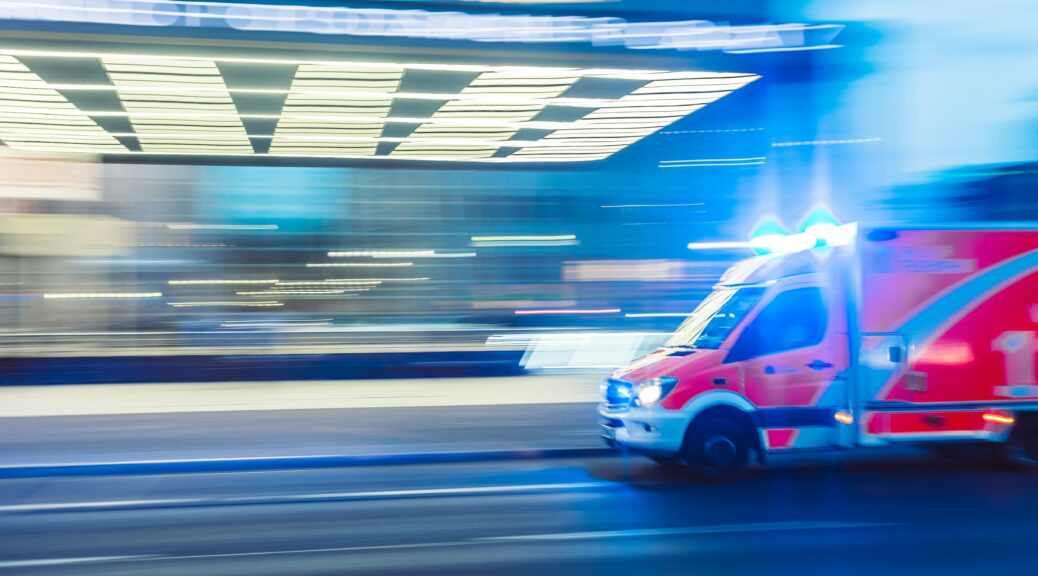 An ambulance on the right side of the screen is driving into a hospital loading zone, but the photographer has moved the camera while taking the photo so single lights look as though they are dragged enhancing the motion-like quality of the photograph and reinforcing the speed at which paramedics must work.