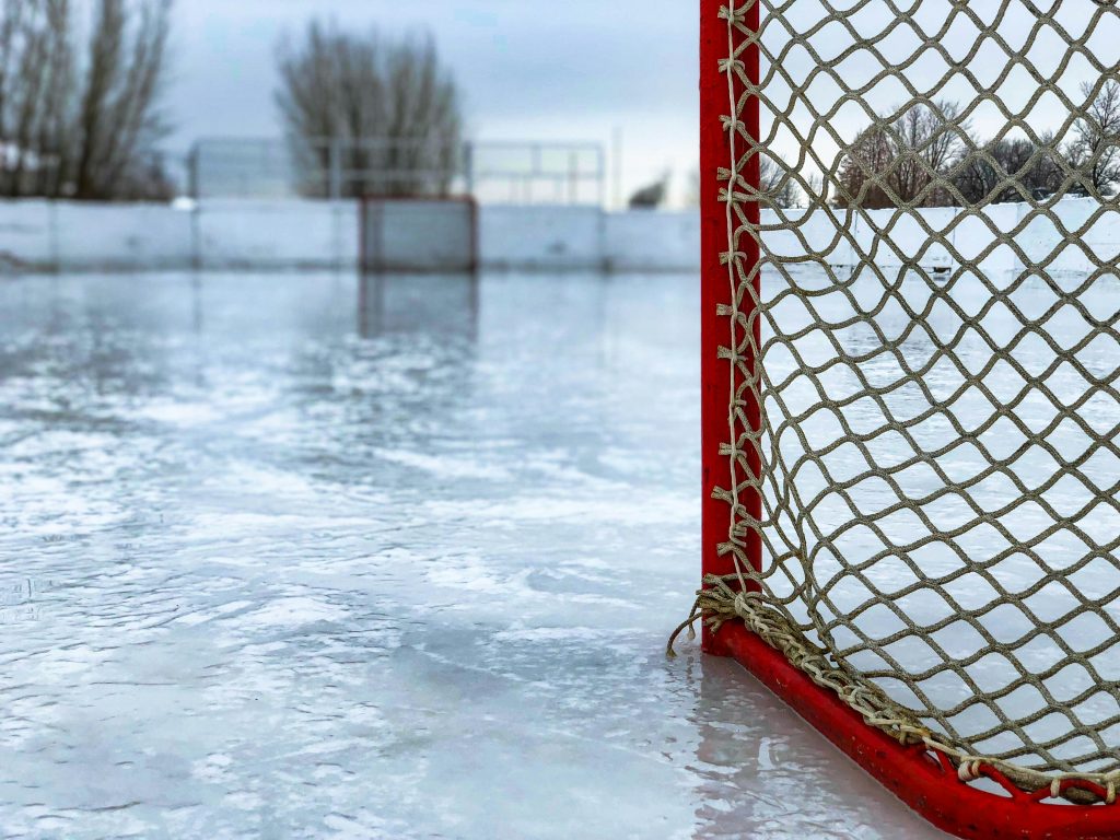 the side of a hockey net with a red frame sits on an empty outdoor ice rink. The net comprises the right side of the photo and the empty rink and trees are blurred in the background