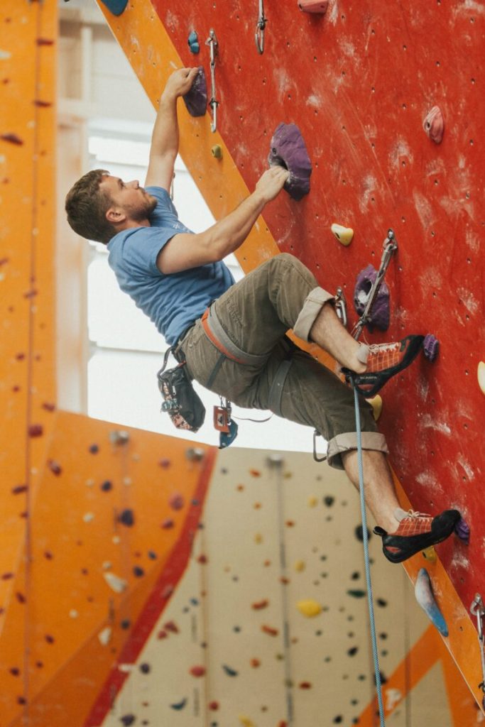 A man in a climbing harness climbs up an orange and red wall in a climbing gym