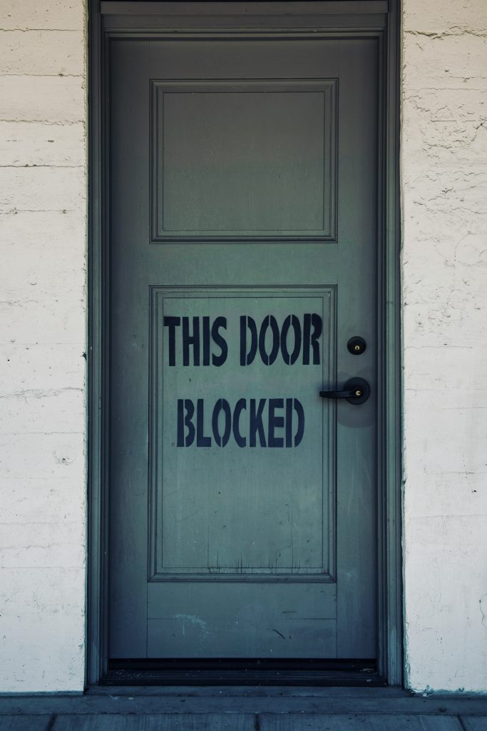 A light green door is inset into a brick wall; in the bottom panel of two on the door, it reads "This Door Blocked" in capital black letters
