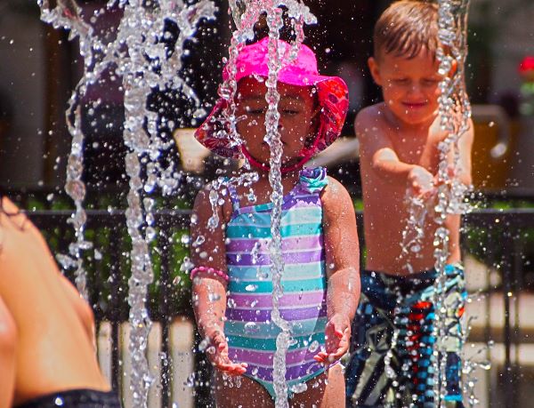 Children play in a water park. Water spouts up as a small girl in a striped pink purple and blue one piece swimsuit and a pink sun hat closely stares and plays. Behind her and on the right on the photo a shirtless bot in blue, white, black and grey Volcom swim trunks catches water from another spout.