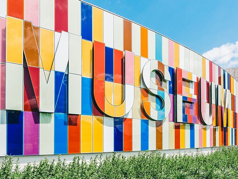 Picture of the front of the Waterloo Region museum. Greenery and grass at the bottom and blue sky above, the multicoloured wall panels with the word "MUSEUM" in multicoloured panels.