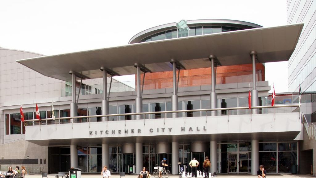 A photo of the front of Kitchener City Hall. In the background, the sky is grey, in the foreground, much of city hall is grey. There are people sitting in front of city hall on chairs and benches. A few people playing giant chess. The words "Kitchener City Hall" are on a veranda above the front doors
