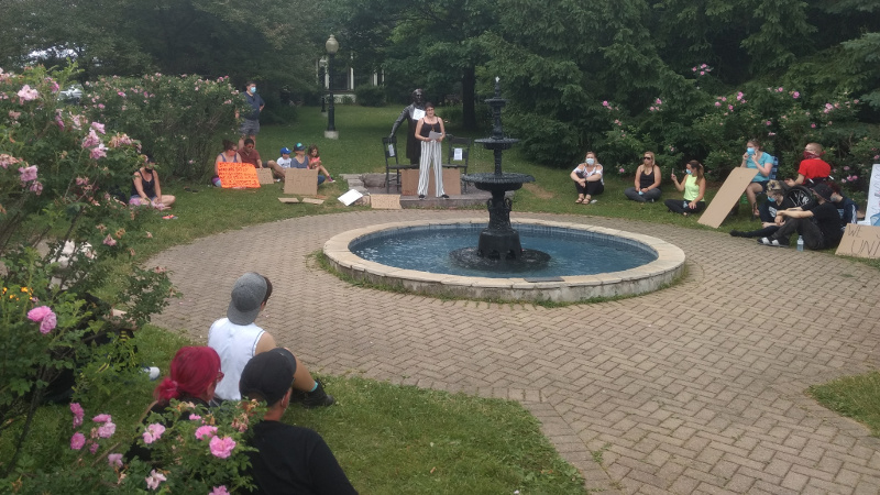 People sitting in the park and listening to Cheyanne Thorpe discuss the negative impacts of the Sir John A MacDonald statue, which stands behind her.