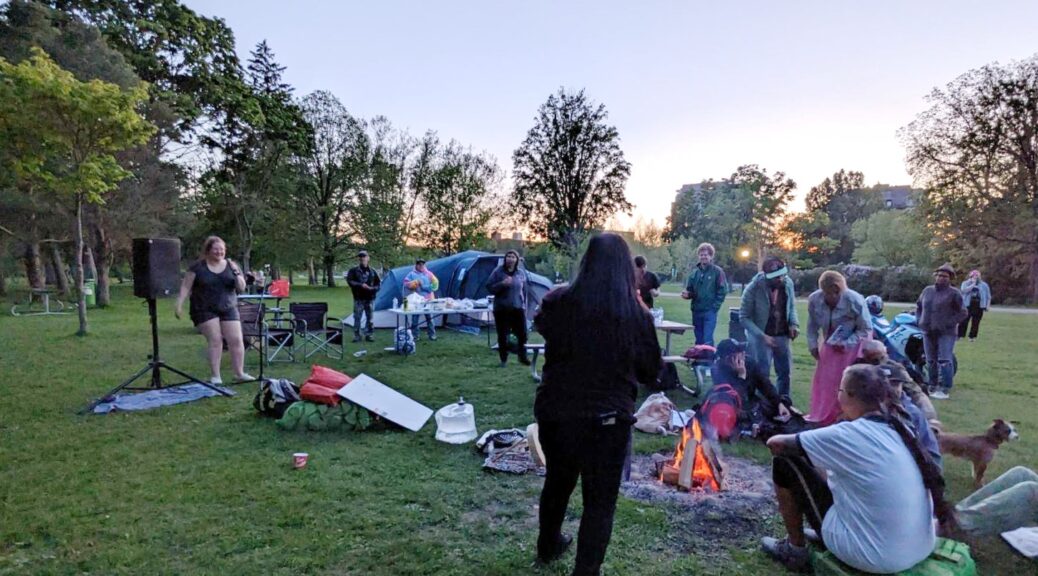 A group of folks in a park with a sunset in the background beyond trees. In the foreground the aforementioned folks with a group of them gathered around a fire. Tents are visible as is the sound system for the event.,