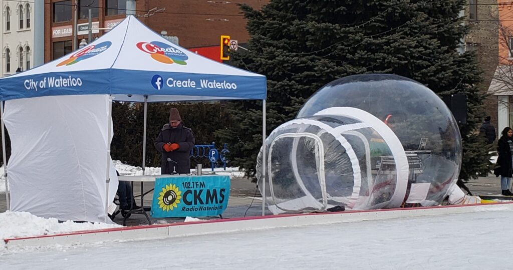 A blue canopy tent with a white wind shield, beside a clear vinyl dome. A man in thick winter closing wearing bright orange gloves is working a table under the canopy. There is a banner on the table with "102.7 CKMS Radio Waterloo.ca"