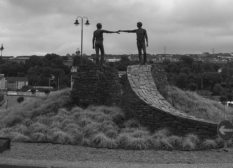 A statue of two life-sized young men each standing on a similar stylised stone structure reaching towards each other. The statue's name is “Hands Across The Divide” by sculptor Maurice Harron and is situated the city of Derry, Northern Ireland