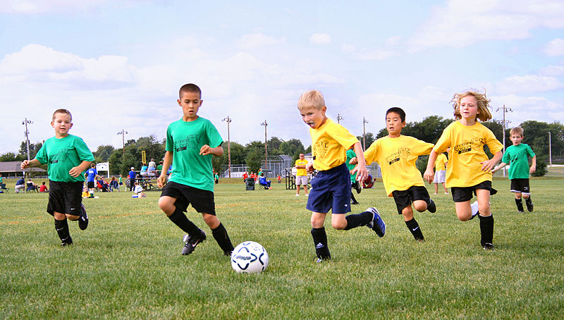 Kids in yellow and green jerseys chase the soccer ball on a green field.