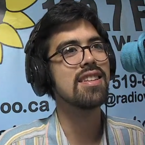 Aaron Liang (a man with dark hair and a Vandyke wearing glasses and headphones; a socked microphone is visible on the right, and a CKMS-FM poster is partially visible in the background)