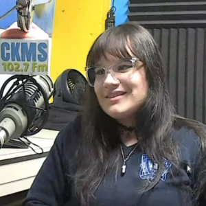 Anabela Tadic (a woman with long dark hair and wearing glasses and a dark sweater sits at at microphone labelled CKMS 102.7 FM)