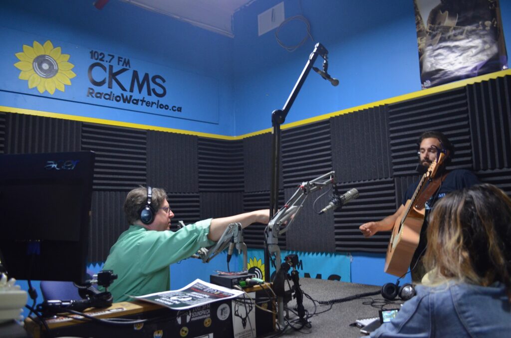 Bob Jonkman wearing headphones and seated at the sound board gestures to Bobcat Bartolo to begin his performance on the air in the CKMS studio.
