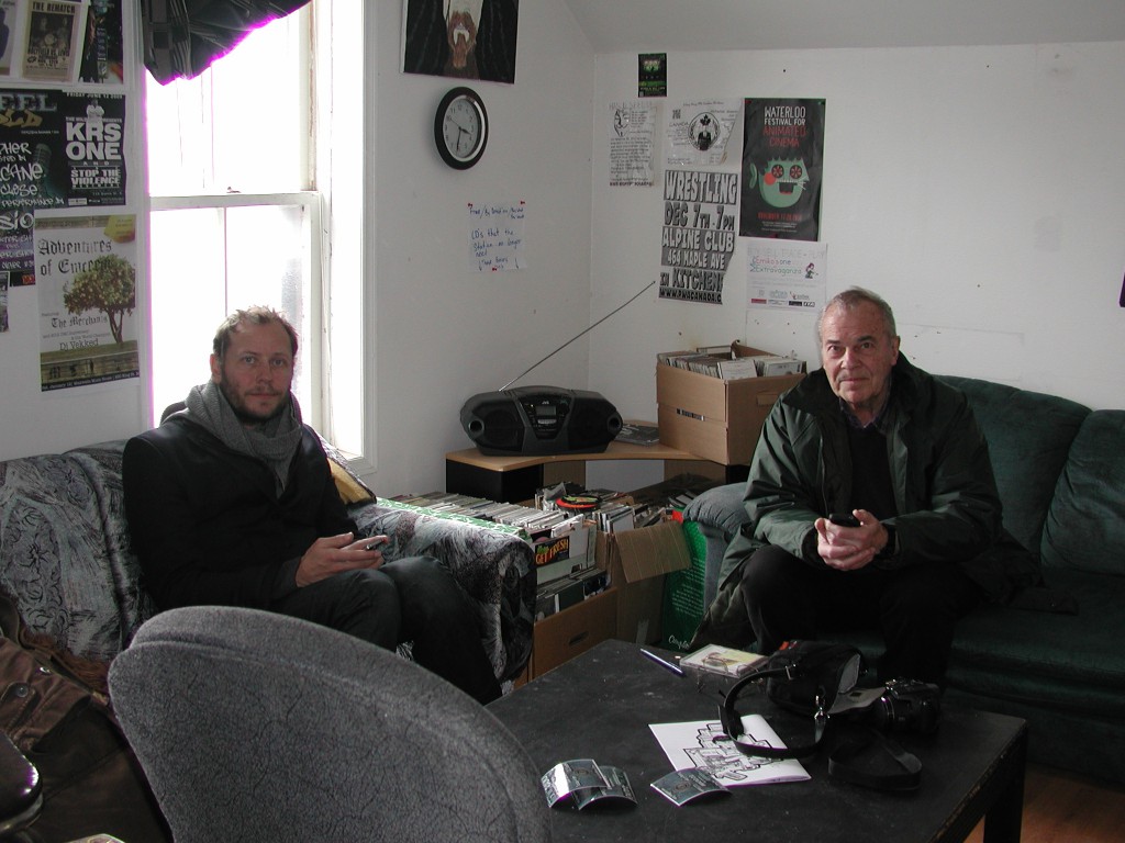 Two men wearing coats sitting indoors on comfy couches
