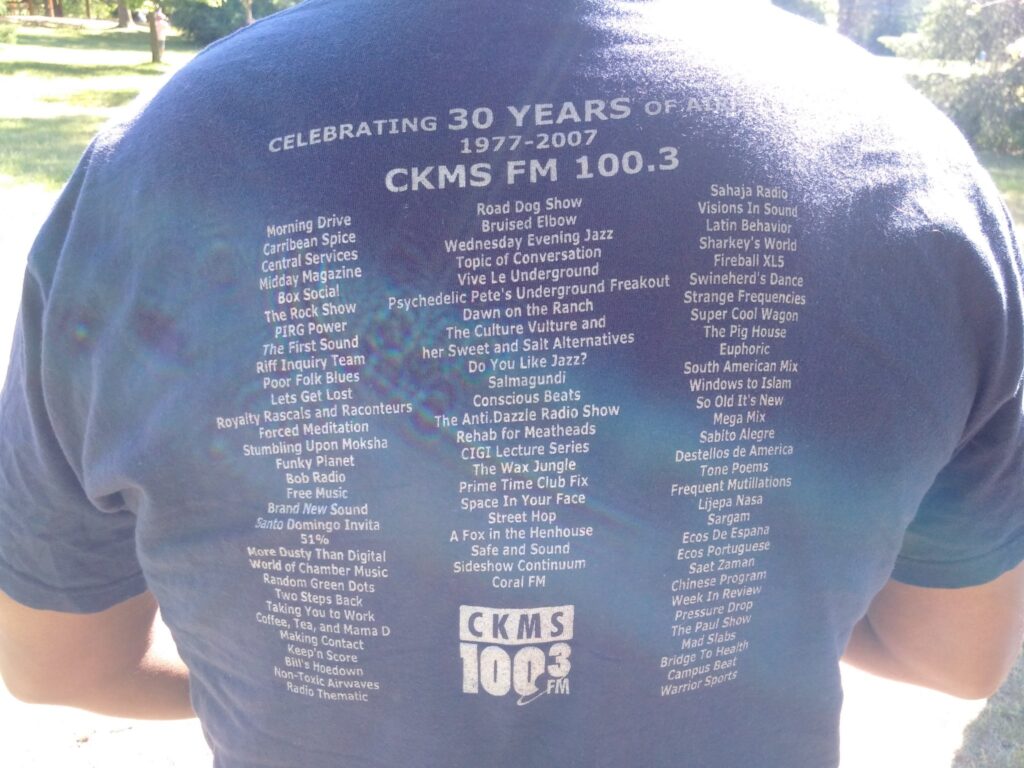 Writing on the back of a T-Shirt for the 30th anniversary of CKMS-FM listing all the shows on CKMS-FM: Celebrating 30 years of air | 1977-2007 | CKMS FM 100.3 | Morning Drive, Carribean Spice, Central Services, Midday Magazine, Box Social, The Rock Show, PIRG Power, The First Sound, Riff Inquiry Team, Poor Folk Blues, Lets Get Lost, Royalty Rascals and Raconteurs, Forced Meditation, Stumbling Upon Moksha, Funky Planet, Bob Radio, Free Music, Brand New Sound, Santo Domingo Invila, 51%, More Dusty Than Digital, World of Chamber Music, Random Green Dots, Two Steps Back, Taking You to Work, Coffee Tea and Mama D, Making Contact, Keep'n Score, Bill's Hoedown, Non-Toxic Airwaves, Radio Thematic, Road Dog Show, Bruised Elbow, Wednesday Evening Jazz, Topic of Conversation, Vive Le Underground, Psychedelic Pete's Underground Freakout, Dawn on the Ranch, The Culture Revolution and her Sweet and Salt Alternatives, Do You Like Jazz?, Salmagundi, Concious Beats, The Anti-Dazzle Radio Show, Rehab for Meatheads, CIGI Lecture Series, The Wax Jungle, Prime Time Club Fix, Space In Your Face, Streep Hop, A Fox In The Henhouse, Safe and Sound, Sideshow Continuum, Coral FM, Sahaja Radio, Visions In Sound, Latin Behaviour, Sharkey's World, Fireball XL5, Swineherd's Dance, Strange Frequencies, Super Cool Wagon, The Pig House, Euphoric, South American Mix, Windows to Islam, So Old It's New, Mega Mix, Sabito Alegre, Destellos De America, Tone Poems, Frequent Mutilations, Lijepa Nasa, Sargam, Ecos De Espana, Ecos Portuguese, Saet Zaman, Chinese Program, Week In Review, Pressure Drop, The Paul Show, Mad Stabs, Bridge To Health, Campus Beat, Warrior Sports | CKMS 100.3 FM (logo)