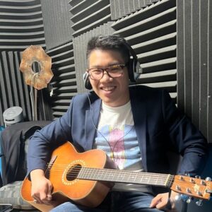 (Caleb Khuu in the CKMS-FM studio wearing headphones and holding a guitar)