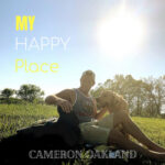 My Happy Place | Cameron Oakland (Cameron Oakland sitting in a field, backlit by the sun, petting two large dogs)