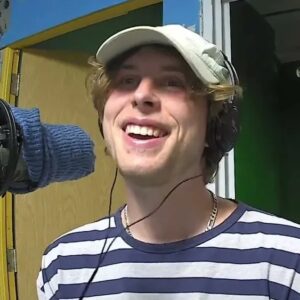 CxViolet (a man with blond hair wearing a striped shirt and headphones over a white baseball cap sits at a microphone covered in a blue sock in the CKMS-FM studio)