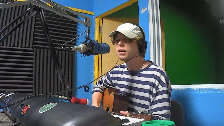 CxViolet, a man wearing a striped shirt and headphones over a white baseball cap sitting at a microphone and plays guitar. There is also a very large synthesizer on the desk in front of him)
