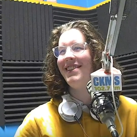 Czar (a person with long hair, glasses, and a yellow shirt, with headphones around their neck sitting at a microphone with mic flag "CKMS 102.7 FM")