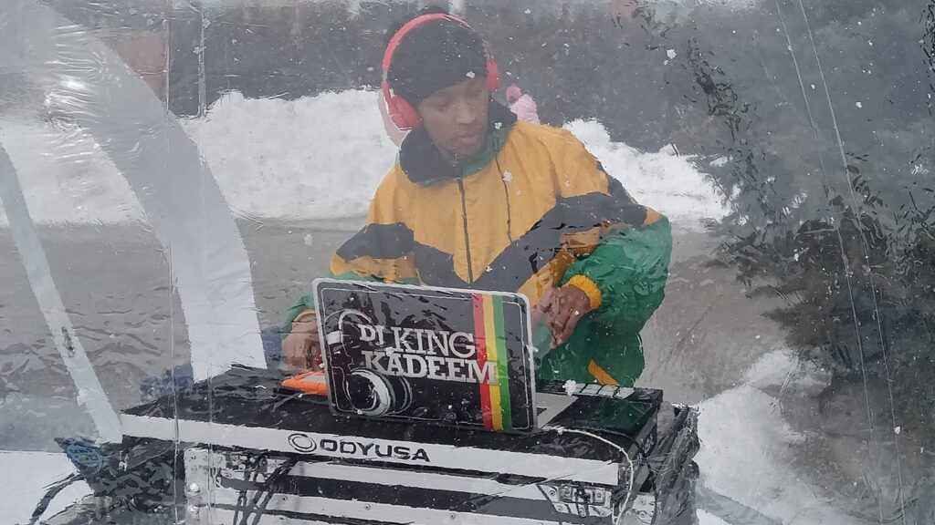 A man wearing red headphones and a bright rasta-coloured jacket standing behind DJ gear, with a sticker labelled "DJ King Kadeem", all inside a clear vinyl bubble.