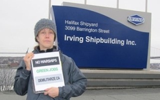 Tamara Lorincz wearing a grey jacket and a grey wool cap holding a poster that reads "No Warships | Green Jobs | Demilitarize.ca" while standing in front of a sign that reads "Irving | Halifax Shipyard | 3099 Barrington Street | Irving Shipbuilding Inc."