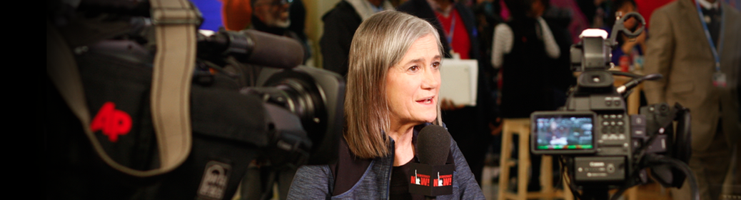 (Amy Goodman at the mic at a press conference surrounded by cameras)
