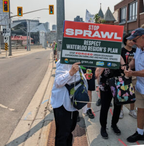A rally participant standing in front of a group with a sign reading "Stop the Sprawl... Respect Waterloo Region's Plan". The setting is on the sidewalk with the empty road on the right or the photo.