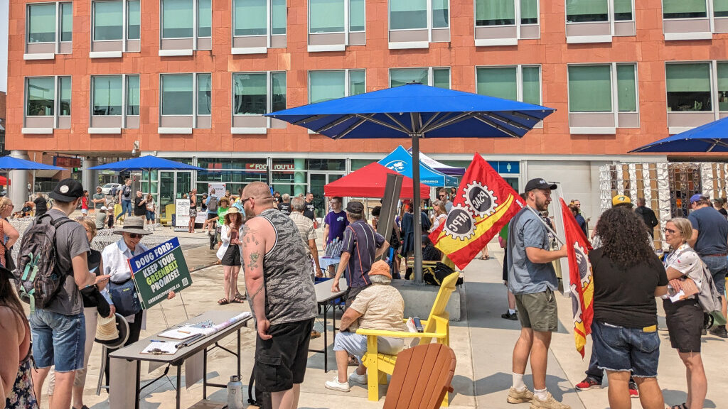 A scene from the Enough is Enough rally in Kitchener. People are milling about, making friends and connections. Organising to take action, declare "Enough is Enough" and take back control of their lives. The background is a wing of Kitchener City hall, and there are blue shade umbrellas providing cover for some of those gathered.