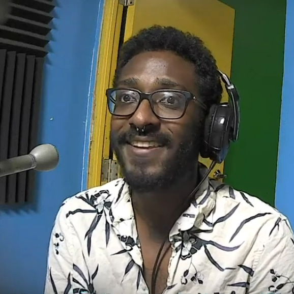 Fabian Fletcher (a man wearing headphones and a white Aloha shirt sits at a microphone)
