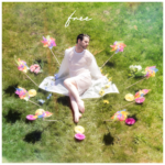 Free (JSJ sitting on a grassy field wearing a white dress, surrounded by flowers)