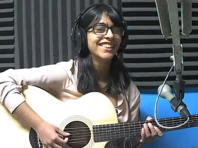 Hiba Farooki (a woman with dark hair and glasses, in front of a microphone, playing guitar)