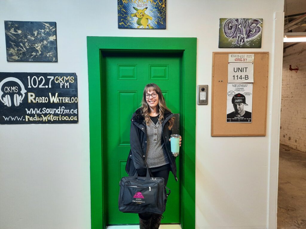 Jody Swannell standing at the studio entrance. A green door with signage CKMS 102.7FM