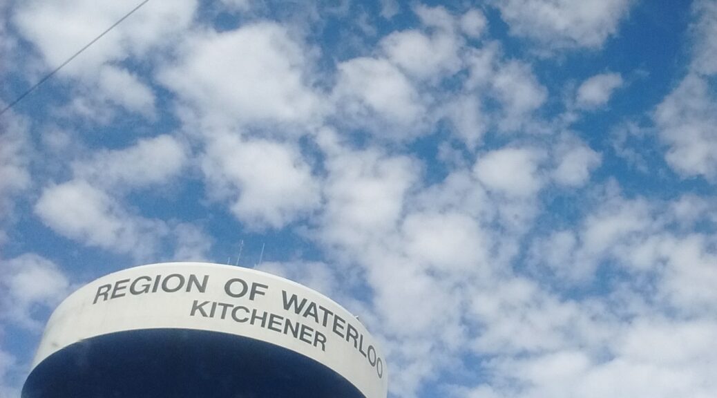 Looking up at the Kitchener's water tower. There are clouds in the sky and on the water tower it reads "REGION OF WATERLOO KITCHENER"