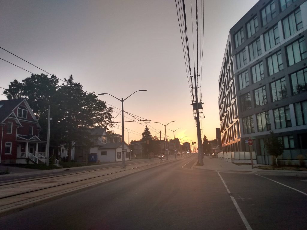 The sun sets over a 5-storey residential building on the right of the image. Cars in the distance drive east beside the LRT line.