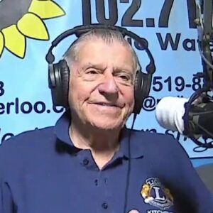 Jack Bishop (a man wearing headphones and a blue golf shirt with a Lions Club logo sits at a microphone in front of a CKMS 102.7 banner)
