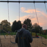 (Man wearing a winter jacket with his back to the camera facing a playground swing set)