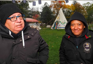 A photo of Shawn Johnston and Amy Smoke wearing toques with the White tee-pee of O:se Kenhionhata:tie/Land back Camp in the background.