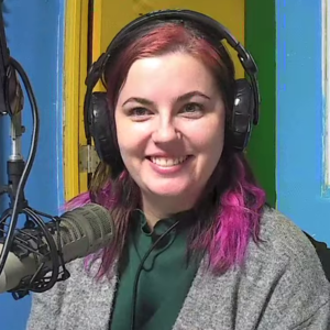 Lillie Proksch (A woman with pink hair wearing a gray cardigan sits in front of a microphone and smiles into the camera)