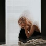 (Paige Warner, a woman with long curly blonde hair sitting in front of white venetian blinds at the bottom right corner of the picture with her head resting on her hands which are on her knees. The photo is multiple exposure, showing faint outlines of Paige lowering her head to her hands. There is a small pile of clothes or blankets beside her)