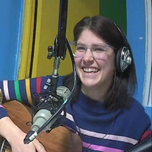 A smiling woman wearing a blue sweater and headphones holds a guitar while sitting in front of a microphone.