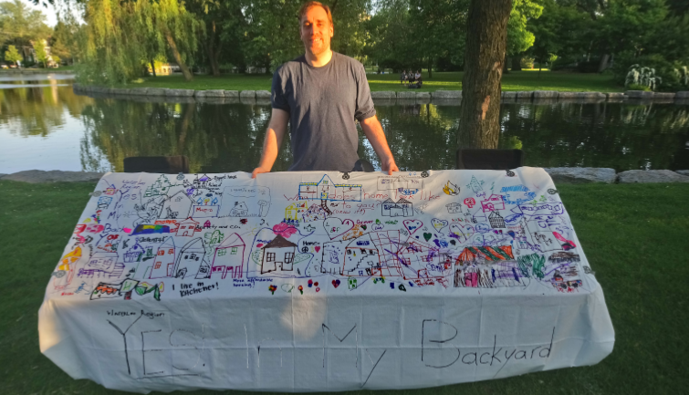 Martin Asling standing behind a banner with homes drawn on it and the words "Yes In My Backyard" in large letters. Trees and a lake are in the background.