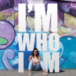 I'm Who I Am (photo of Mary Abdel-Malek Neil sitting in front of a wall covered in large graffiti)