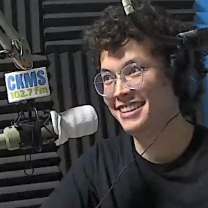 Matt Lam (a man with dark curly hair and glasses wearing headphones in front of a microphone with a flag "CKMS 102.7 FM", the background is black acoustic tile)