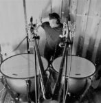 (Black and white photo of Mistahi Corgil sitting at tympani drums with mics on mic stands in the foreground)